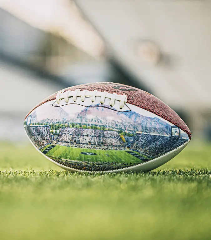 A photograph of a football featuring a picture of the BYU stadium on its side sitting on turf