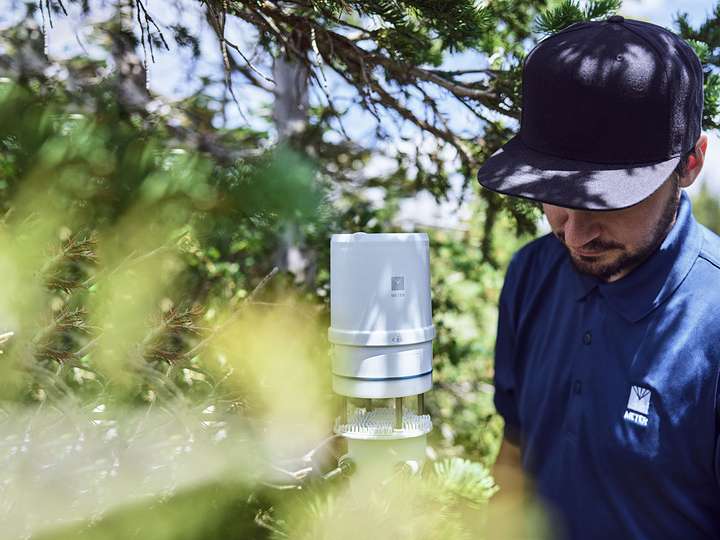 A photograph of a researcher in the field checking on a mounted ATMOS 41 in a stand of trees