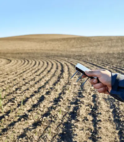 A researcher's hand holding a TEROS 12 soil moisture sensor over a plowed field